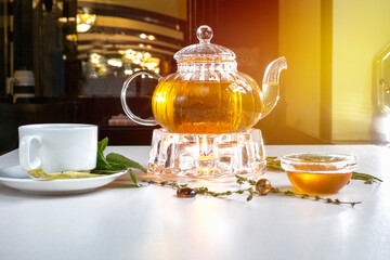 Fragrant fresh herbal tea and melissa in a glass teapot warming on a candle on a white table. Traditional herbal drink. Tea with honey. Kettle warmer