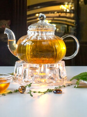 Obraz na płótnie Canvas Fragrant fresh herbal tea and melissa in a glass teapot warming on a candle on a white table. Traditional herbal drink. Tea with honey. Kettle warmer