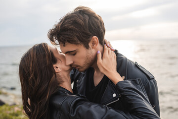 Fototapeta premium Young woman touching neck of boyfriend in leather jacket on beach