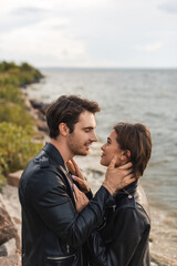 Obraz premium Side view of young couple in leather jackets touching and looking at each other near sea