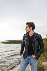 Young man in jeans and leather jacket looking away on sea coast