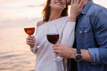 Cropped view of woman touching boyfriend while holding glass of wine near sea at sunset
