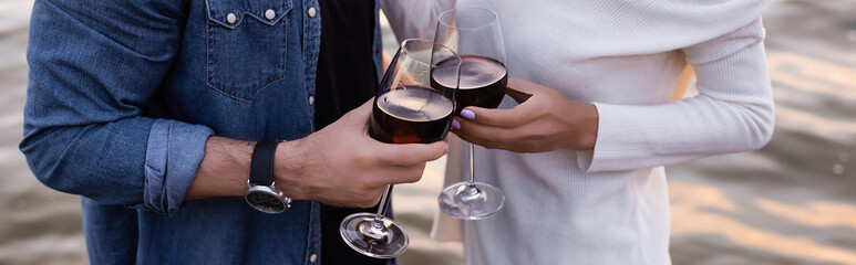 Horizontal crop of couple holding glasses of wine near sea