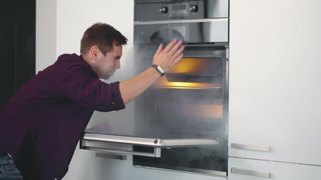 Young Shocked Man Opening Oven Filled With Smoke In Kitchen