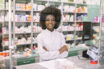 Fototapeta premium Happy young dark skinned woman pharmacist, posing to camera with smile, standing with arms crossed over drugstore background. Medicine, pharmacy, people, health care and pharmacology concept