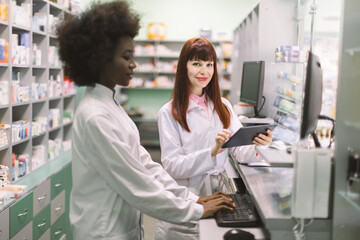 Obraz premium Two young cheerful pharmacists women working together at pharmacy. African woman typing on computer while her Caucasian colleague using tablet. Focus on Caucasian woman