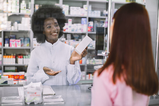 Pharmacy, People And Healthcare Concept. Smiling Female Afrcian Pharmacist Giving Medicine To Young Caucasian Woman In Drugstore. View Through The Patient Shoulder