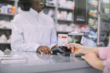 Cropped image of smiling African woman pharmacist holding terminal and hands of Caucasian woman patient, paying for medicines with credit card in pharmacy