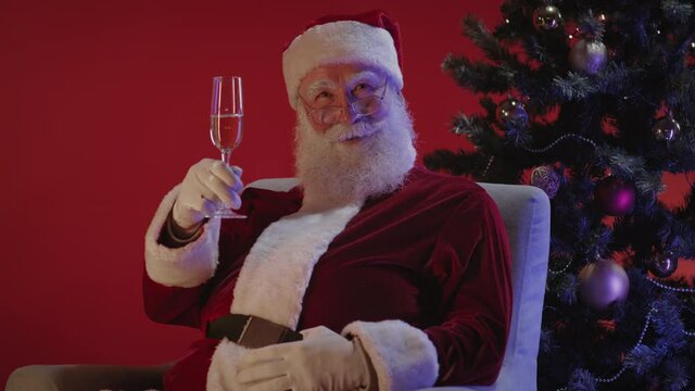 Medium shot of Santa Claus sitting on armchair standing near Christmas tree, congratulating on New Year and Merry Christmas and then making sip of champagne