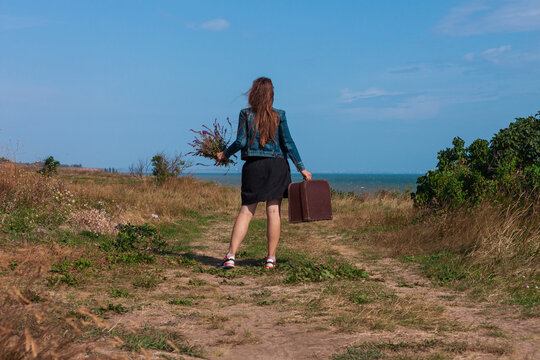 Young woman with long brown hair in denim jacket, black skirt, vintage suitcase,purple wild flowers bouquet off-road sea landscape. Hitchhiker on countryside trip. Lifestyle photo lady walking outdoor