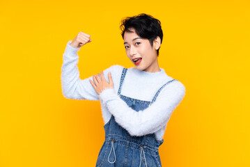Young Asian girl in overalls over isolated yellow background making strong gesture