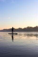 Teenager boy rowing on SUP (stand up paddle board) in autumn Danube river at foggy morning against...