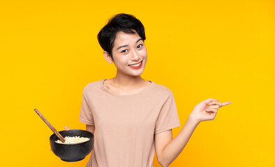 Young Asian girl over isolated yellow background surprised and pointing finger to the side while holding a bowl of noodles with chopsticks