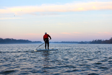 Back view on man rowing on SUP (stand up paddle board) in autumn Danube river at foggy morning against the backdrop of autumn trees without leaves