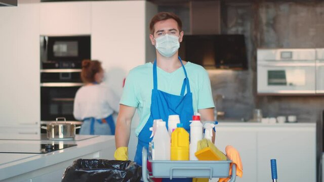 Portrait of male cleaner in uniform and safety mask standing near cart with detergents in kitchen