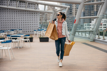 Smiling young woman holding coffee cup, walking with grocery shopping paper bag with long white...