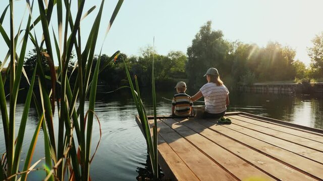 Rear view of a mother with his little son are fishing while sitting on a wooden pier by the lake on a sunny summer day