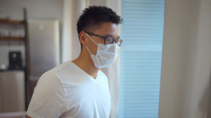 Young asian man wearing safety mask looking out window at home 
