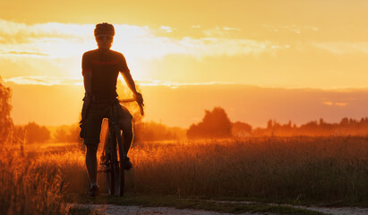 Cyclist on a gravel bike stands in a field on a dramatic sunset background. Beautiful landscape of...