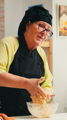 Mature experienced baker recording culinary recipe tutorial using modern camera in kitchen wearing bonete and apron. Influencer chef using internet technology communicating on social media.