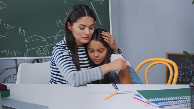 mother becalming and hugging upset schoolkid near desk with notebooks