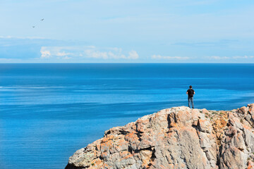 Fototapeta na wymiar Man standing on a rock and admiring beautiful view on the Baikal Lake – the largest freshwater lake in the world. Travel concept. Majestic landscape of Siberian Lake. Image for card or poster.
