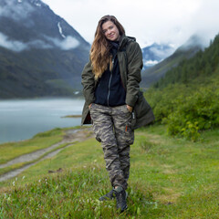 Naklejka premium Young woman in a jacket and trousers against the backdrop of mountains. Girl tourist in warm clothes at dawn against the background of fog, lake blue mountains.