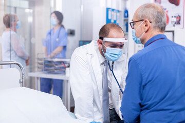 Fototapeta premium Medical practitioner checking heart beat of senior patient during consultation with stethoscope and wearing face mask againts covid-19. Medical examination for infections, disease and diagnosis.