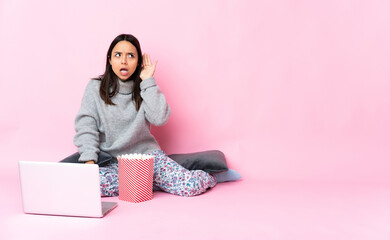 Young mixed race woman eating popcorn while watching a movie on the laptop listening to something by putting hand on the ear