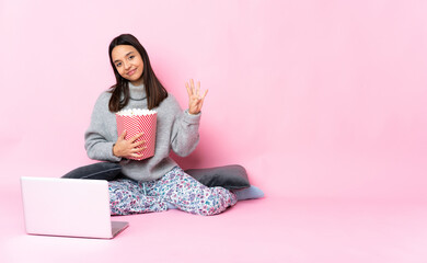 Obraz na płótnie Canvas Young mixed race woman eating popcorn while watching a movie on the laptop happy and counting four with fingers