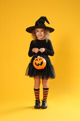Cute little girl with pumpkin candy bucket wearing Halloween costume on yellow background