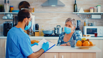Doctor with face mask discussing about coronavirus pandemic with senior woman during home visit and taking notes. Male nurse social worker at retired senior couple visit explaining covid-19 spreading