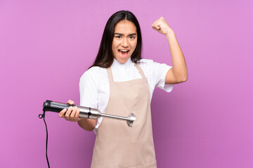 Indian woman using hand blender isolated on purple background making strong gesture