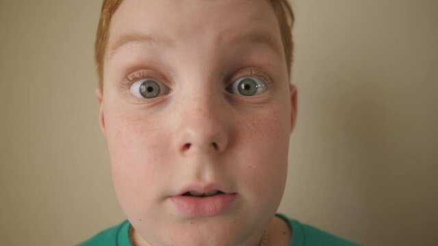 Portrait of ginger kid with freckles showing fright indoor. Little red-haired boy looking into camera with shocked expression inside. Close up scared male face of small child. Slow motion