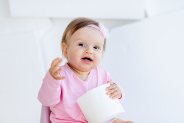 a little girl sits on a potty in pink clothes in a bright room and plays with toilet paper