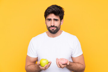 Young handsome man with beard over isolated yellow background taking a chocolate tablet in one hand...