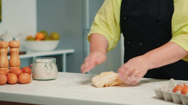 Close up of elderly woman chef hands kneading dough on table in modern kitchen. Retired elderly baker with bonete mixing ingredients with wheat flour preparing for baking traditional cake and bread