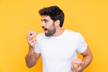 Young handsome man with beard over isolated yellow background eating a donut