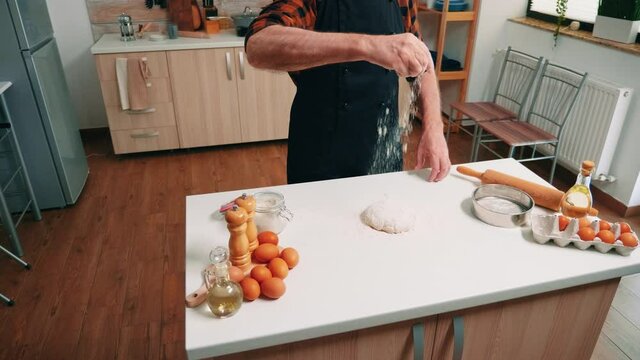 Bread preparation at home in the kitchen by old man. Elderly senior baker with bonete and uniform sprinkling, sifting, spreading rew ingredients on dough by hand, baking homemade pizza and cookies