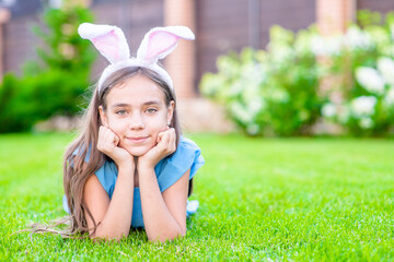 Joyful young girl wearing bunny ears on Easter day lies on green summer grass and looks at camera. Empty space for text