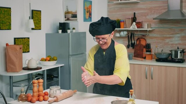 Elderly chef standing in kitchen at home and cooking with enjoyment bakery products of flour. Professional cook claping and powder fly sprinkling rew ingredients over dough preparing traditional bread