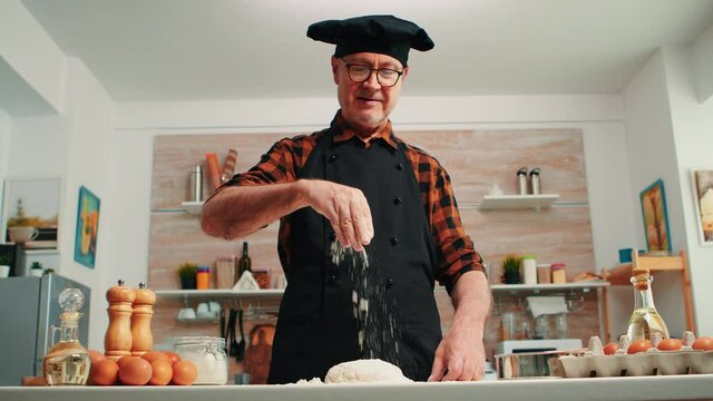 Senior man adding flour on dough by hand looking at camera smiling. Retired elderly chef with bonete and uniform sprinkling, sieving spreading rew ingredients with hand baking homemade pizza and bread