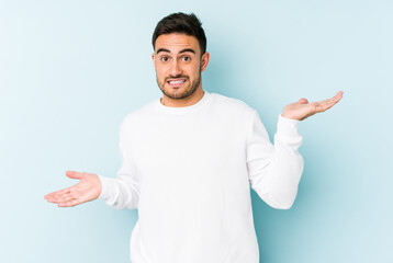 Young caucasian man isolated on blue background confused and doubtful shrugging shoulders to hold a copy space. - obrazy, fototapety, plakaty
