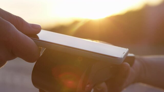 SLOW MOTION, CLOSE UP, LENS FLARE, DOF: Unrecognizable person uses their smartphone to pay for their purchase. Person touches the card reader with their cell phone to process payment on sunny evening.