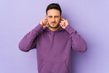 Young caucasian man isolated on purple background covering ears with fingers, stressed and desperate by a loudly ambient.