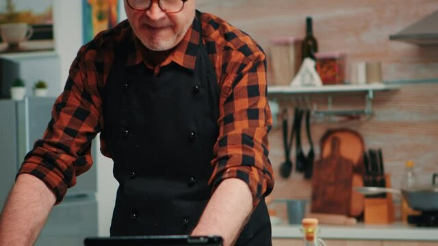 Senior man with bonete and kitchen apron using tablet preparing home cakes for nephews. Retired man following culinary advice on laptop, learning cooking tutorial on social media, forming the dough