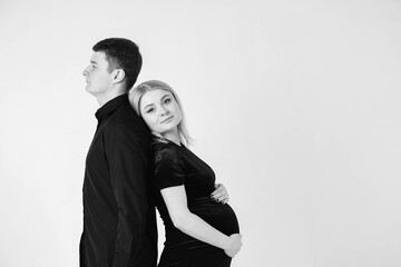 Pregnant woman and her husband smiling in black clothes on a white background. Black and white picture. Beautiful married couple. Copy, empty space for text