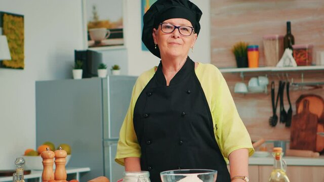 Old lady wearing chef apron and bonete in home kitchen with flour on table for baking looking at camera and smile. Retired elderly baker preparing bakery ingredients to cook homemade bread and cakes