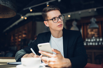 Serious cafe administrator with smartphone writing in copybook