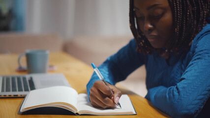 Close up of african beautiful young woman using laptop computer writing notes in notebook studying.